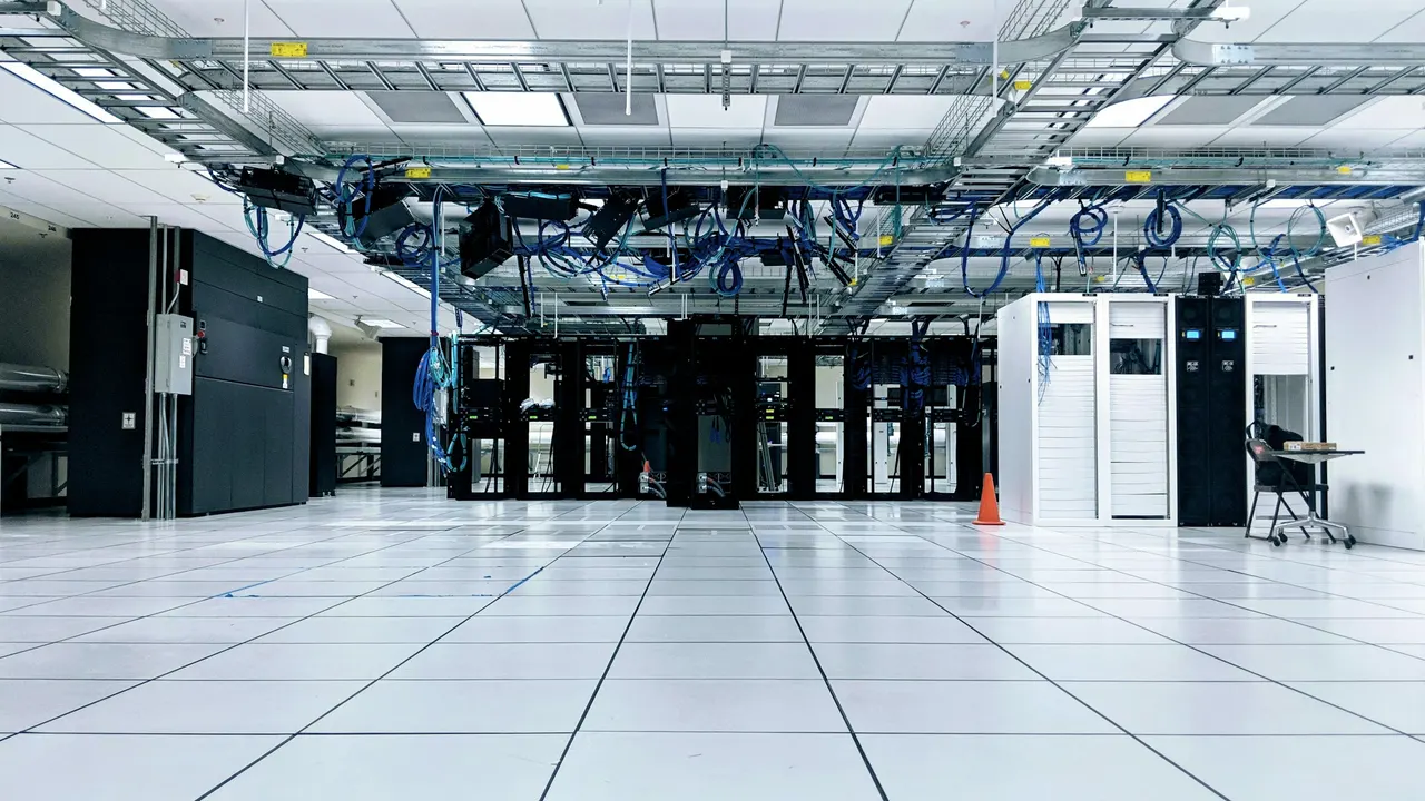 A bright white data hall, rows of black cabinets under cable trays and a raised tile floor